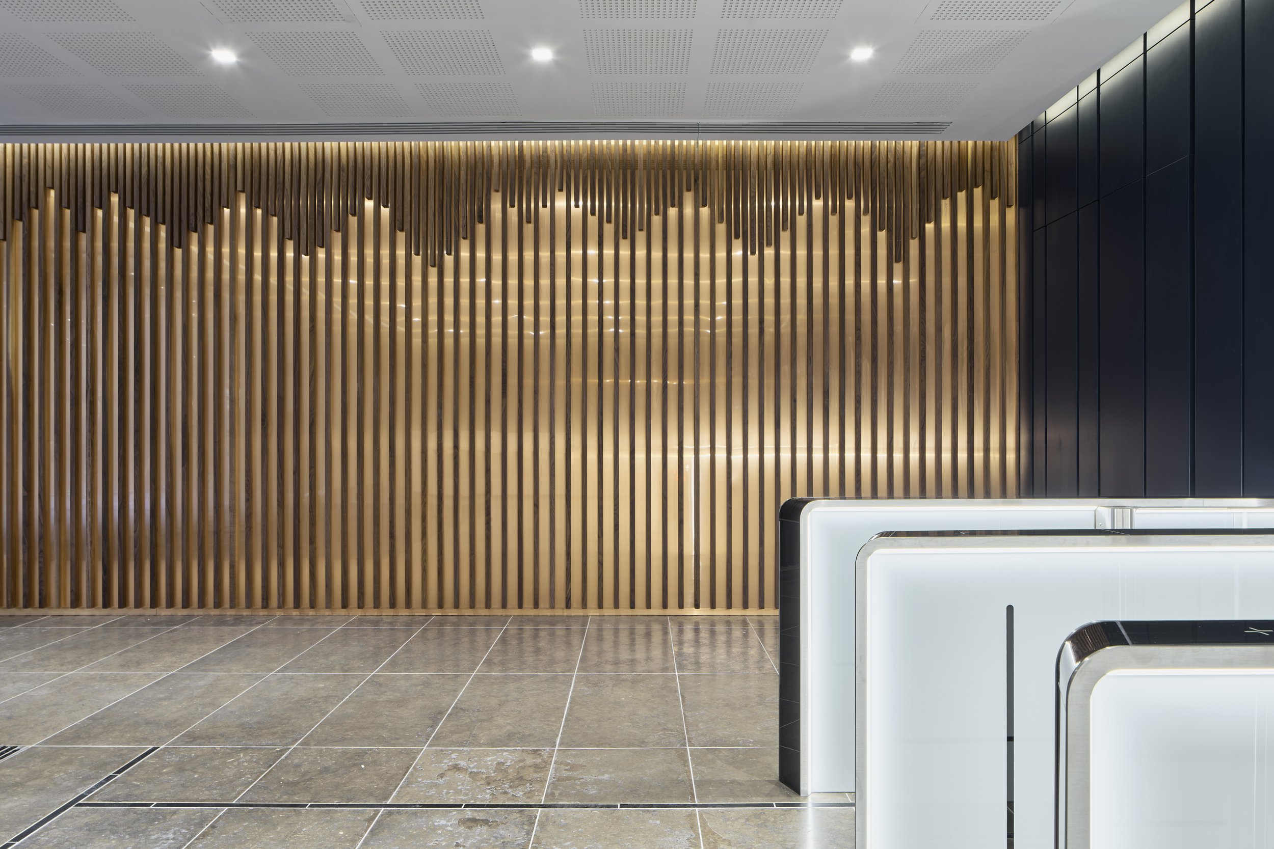 Modern reception area with lighted white curved desks, a wooden slat accent wall, and dark paneling, illuminated by ceiling lights.