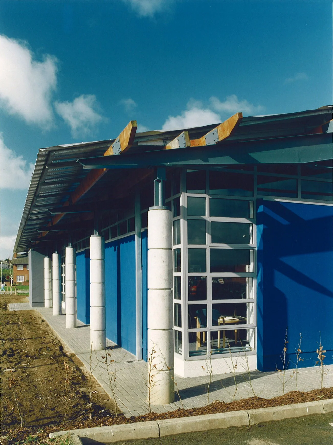 The image shows a modern building with a blue exterior wall, large glass windows, and a sloped metal roof under construction. The area surrounding the building has some small plants and dirt patches.