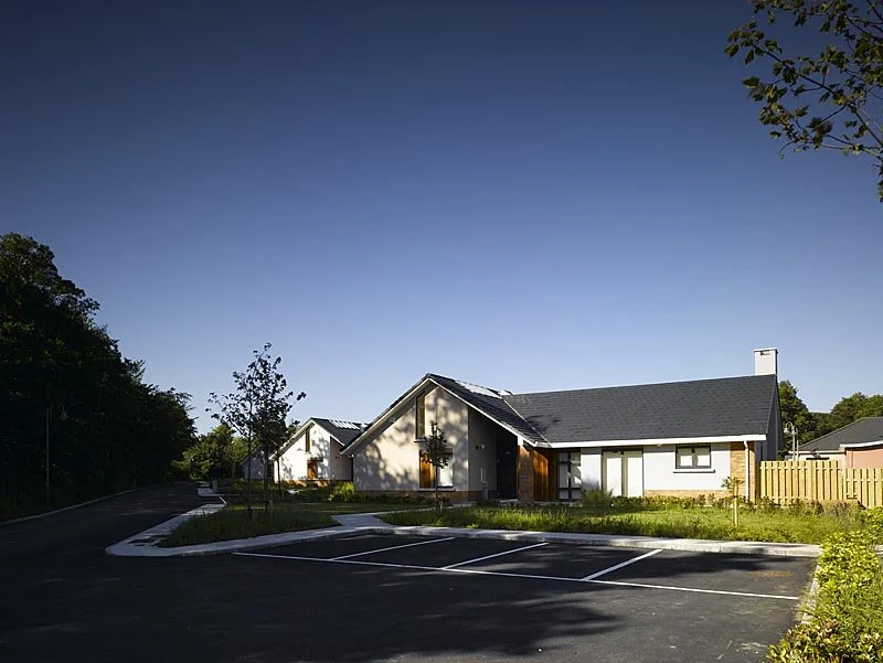 A modern residential house with a dark gray roof, white walls, and wooden accents, situated in a suburban neighborhood with a parking lot in the foreground and trees around.
