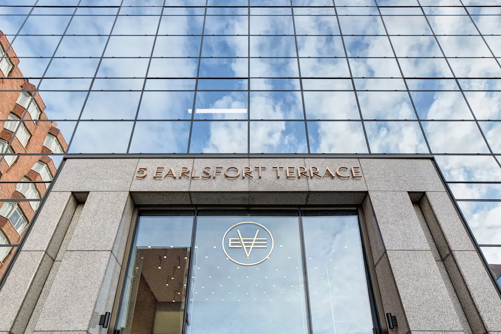 The exterior of a modern glass building with the sign '5 EARLSFORT TERRACE' above the entrance and the logo of a circle with vertical and horizontal lines inside, reflecting the blue sky with clouds.