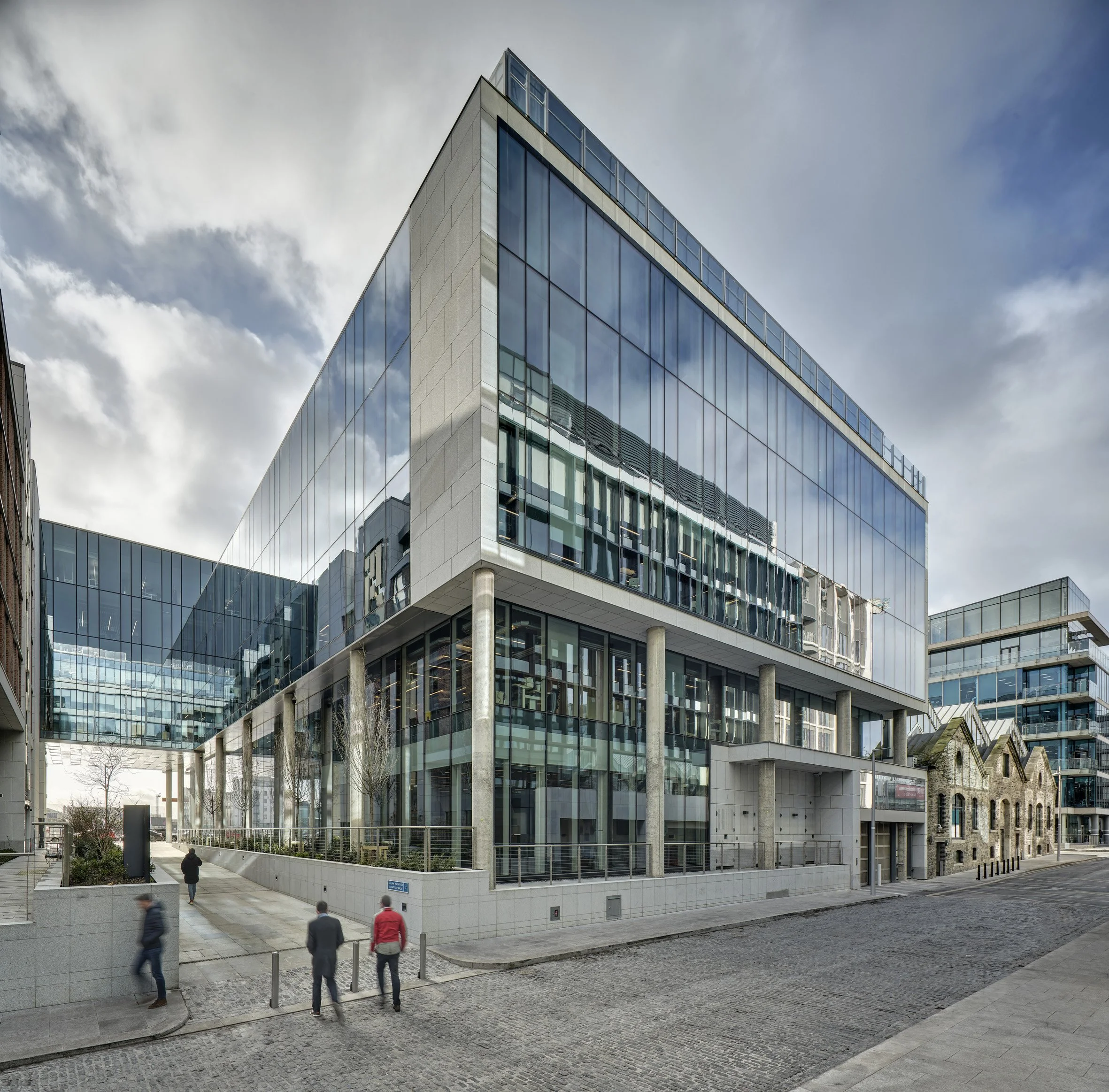 Modern glass office building with reflections of other buildings, and a pedestrian walkway with people walking.