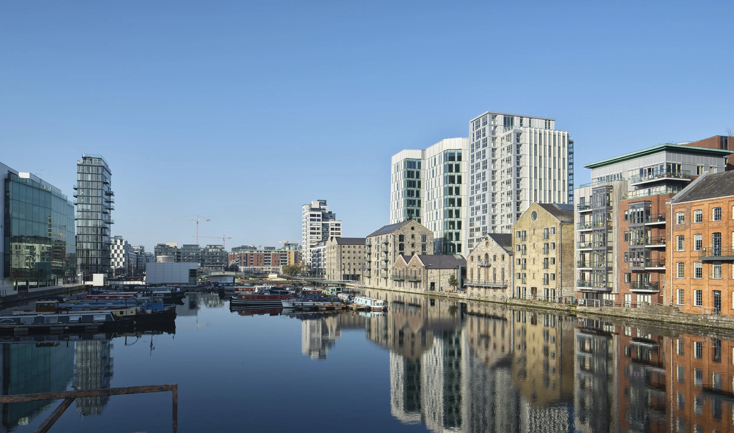 Urban waterfront with modern and historic buildings, boats moored in a canal, and clear blue sky.