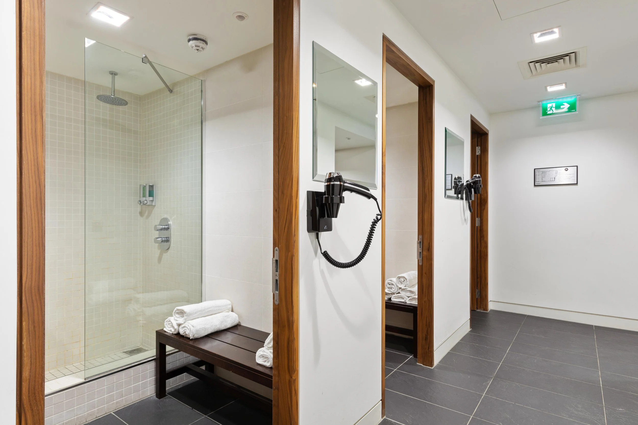 A hotel bathroom hallway with two shower stalls, each with a bench and towels, and mounted hairdryers on the wall.