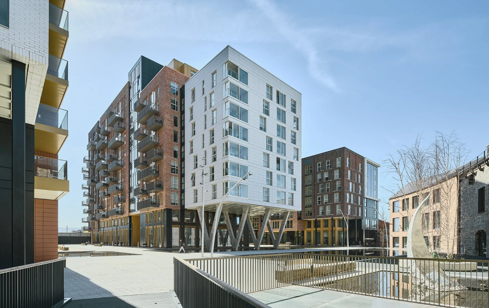 Modern multi-story apartment buildings with balconies, a small water feature, leafless trees, and decorative sculptures in an urban area with a pedestrian walking by.