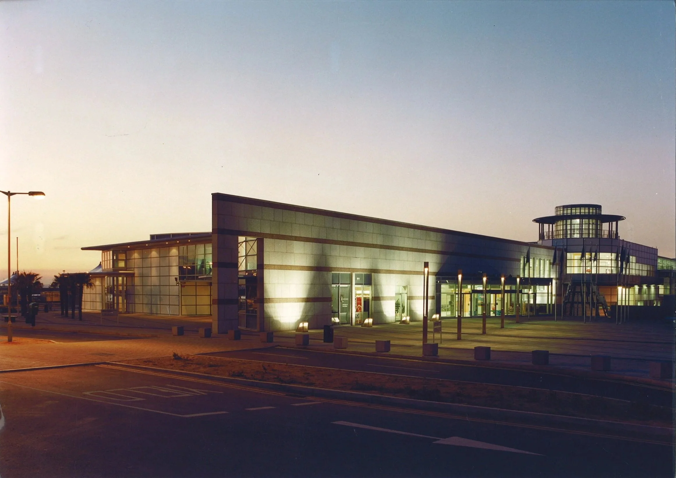 Modern building with large glass windows and a cylindrical tower, illuminated at dusk or dawn, with a parking lot in the foreground.