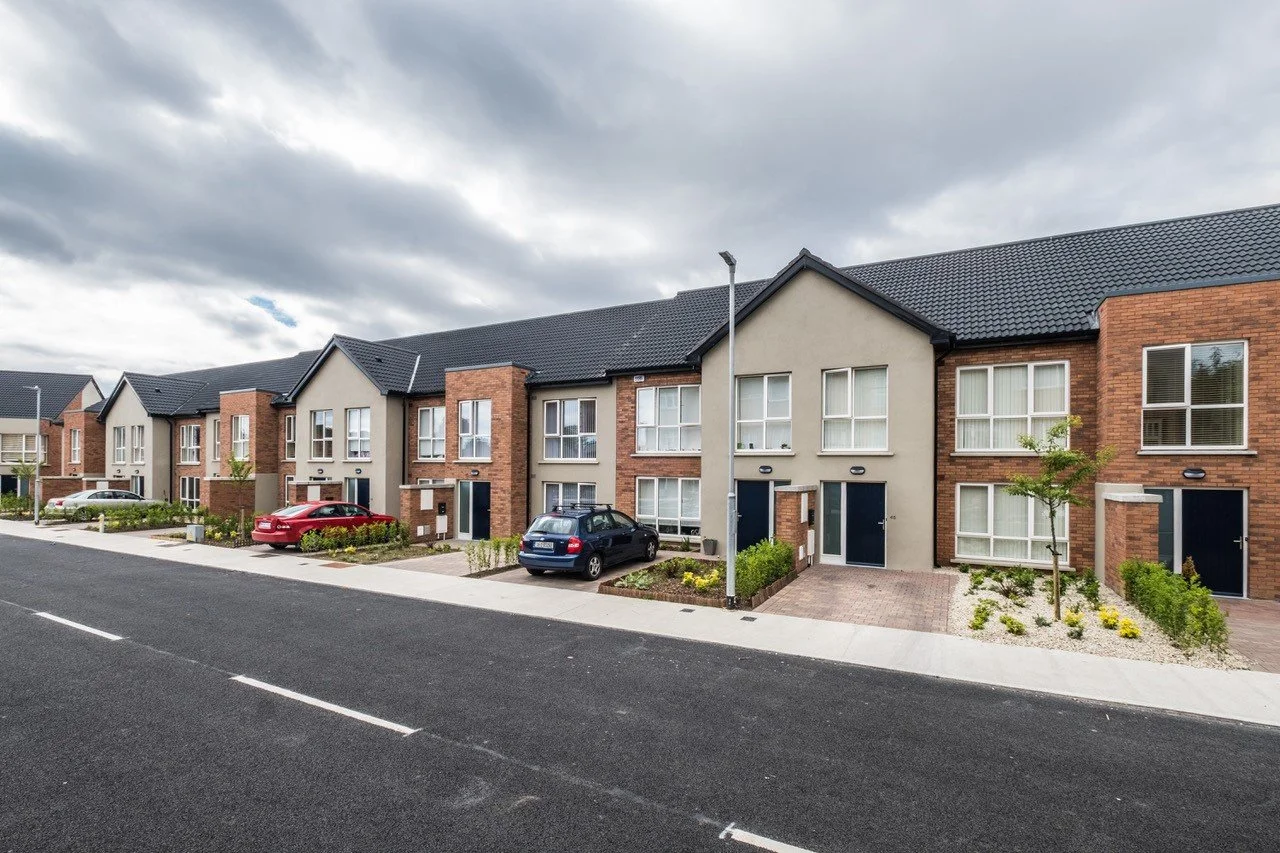 Row of modern townhouses with brick and beige exteriors, black roofs, parked cars, and small front yards, under a cloudy sky.