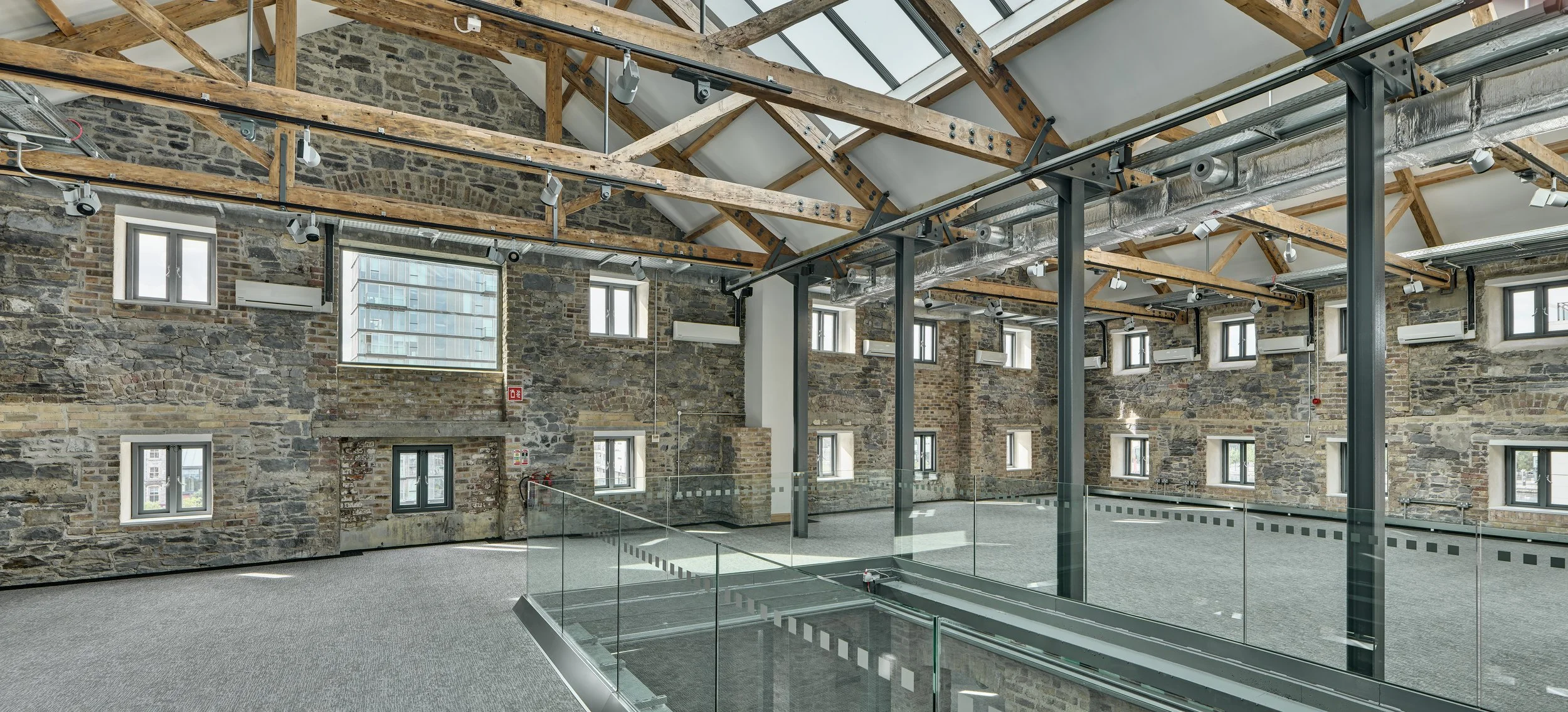 An empty, modern industrial-style interior with exposed brick walls, wooden ceiling beams, multiple small windows, and a glass railing overlooking a lower level.