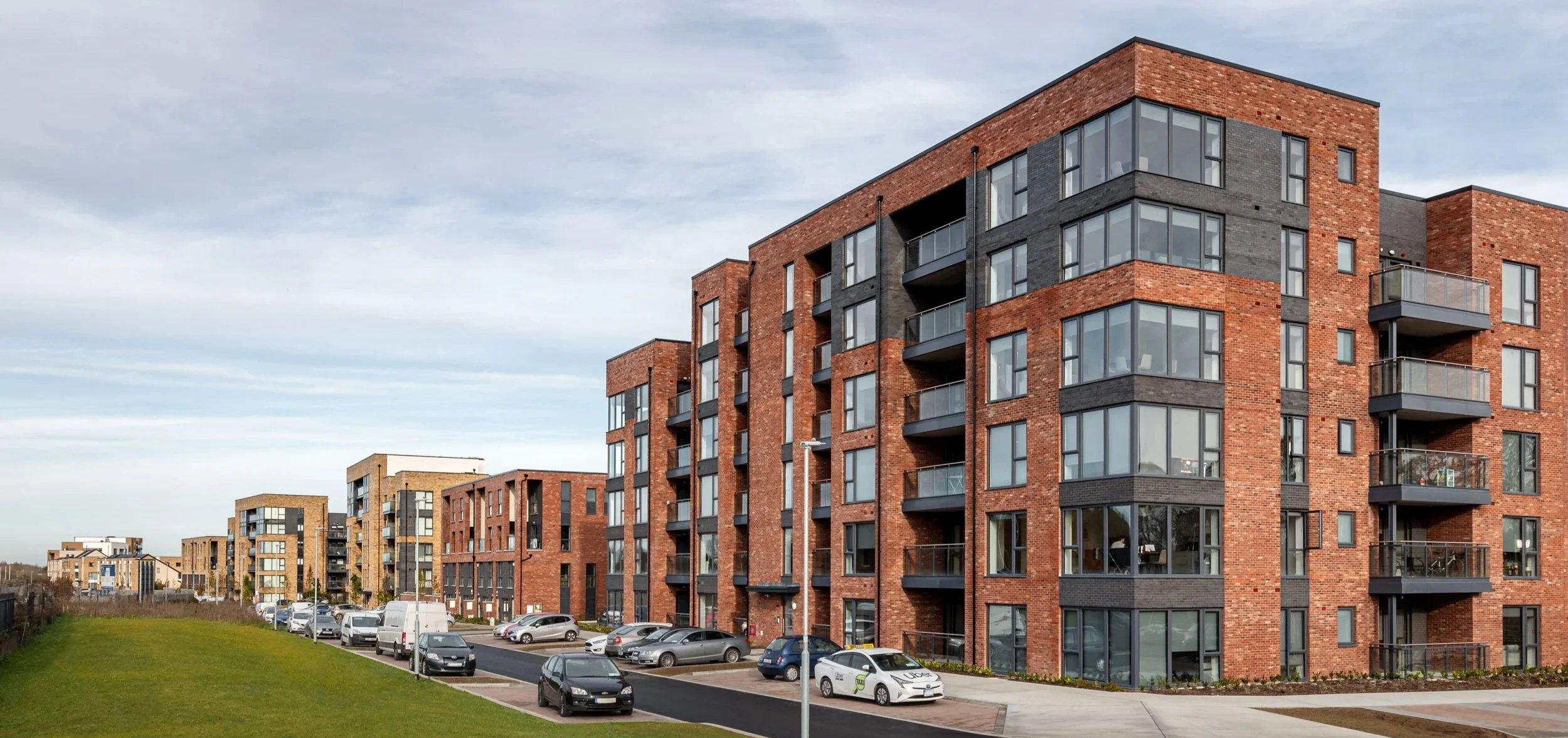 Modern multi-story apartment buildings made of red and gray bricks with large windows and small balconies, parking lot in front with cars, and a grassy area on the side under a cloudy sky.