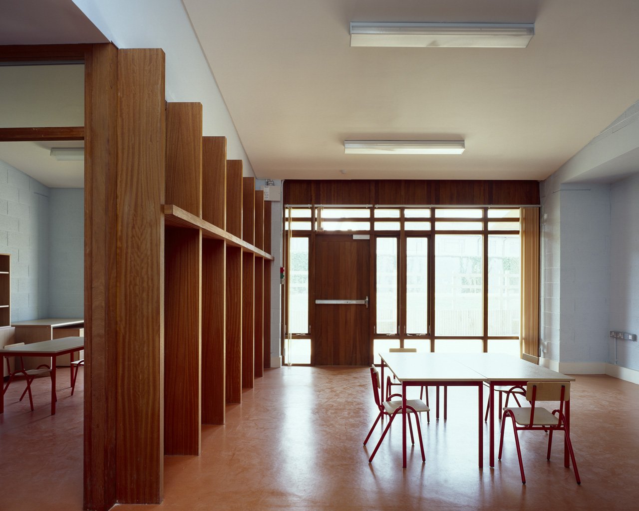 Empty classroom with wooden shelving, tables, and chairs, illuminated by natural light from large windows and glass door.