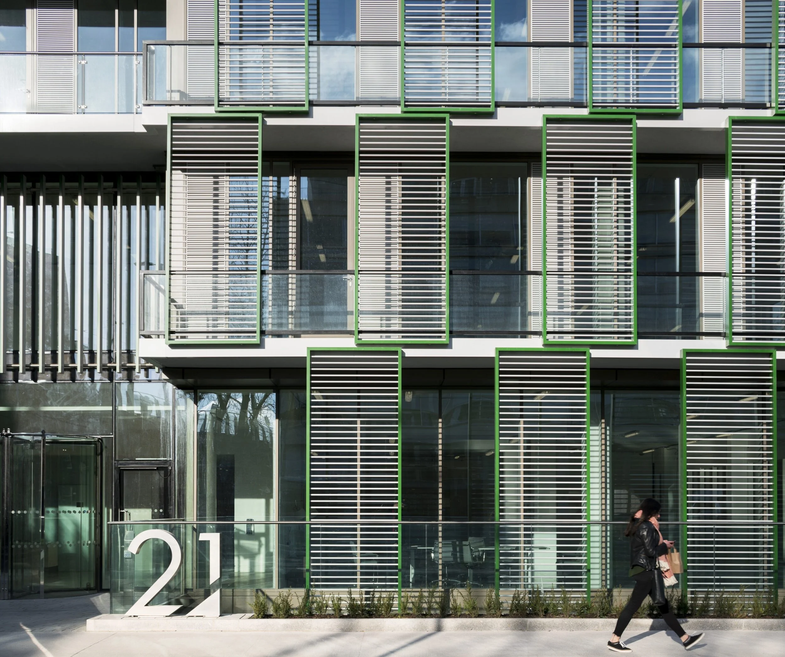 Modern building with glass and metal exterior, green accents, large number '21' sign near the entrance, and a person walking past with a shopping bag.