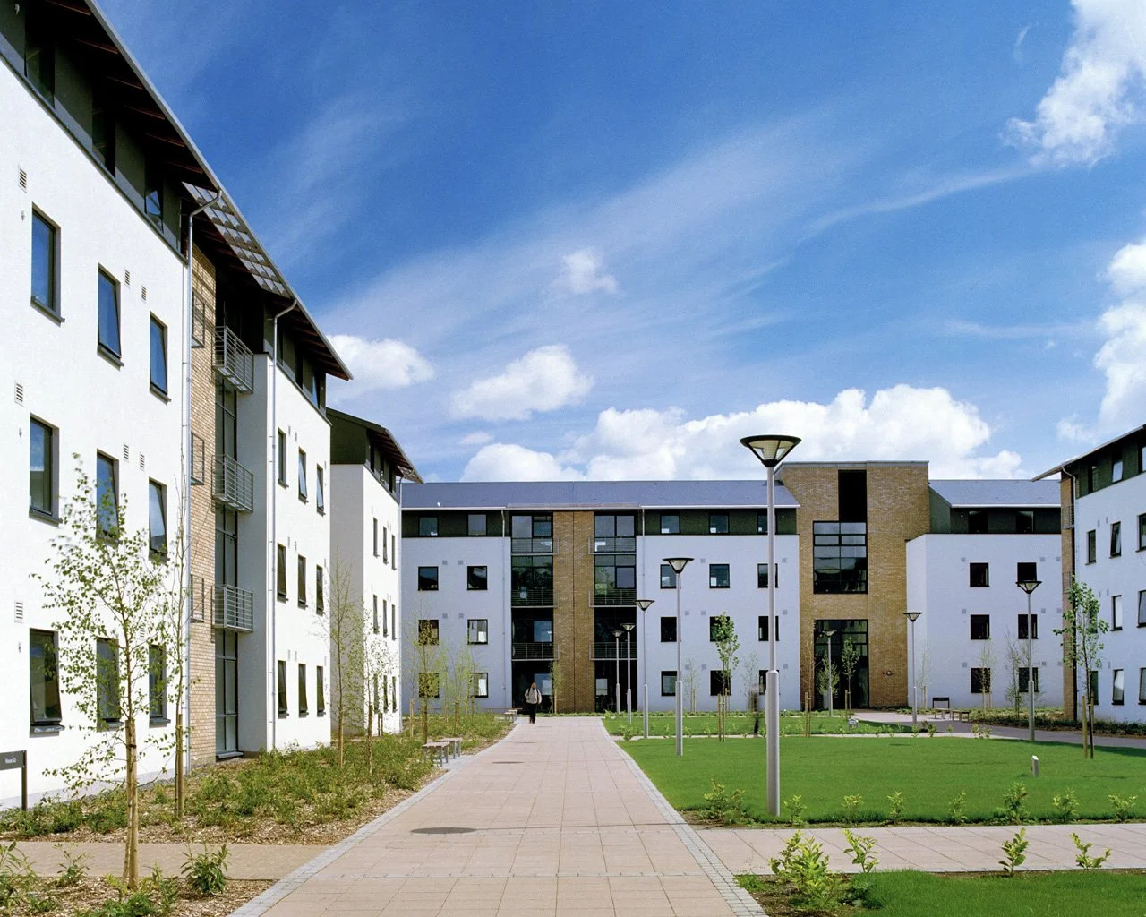 Modern apartment complex with white and brown buildings, a paved walkway, green lawns, young trees, and streetlights under a blue sky with clouds.