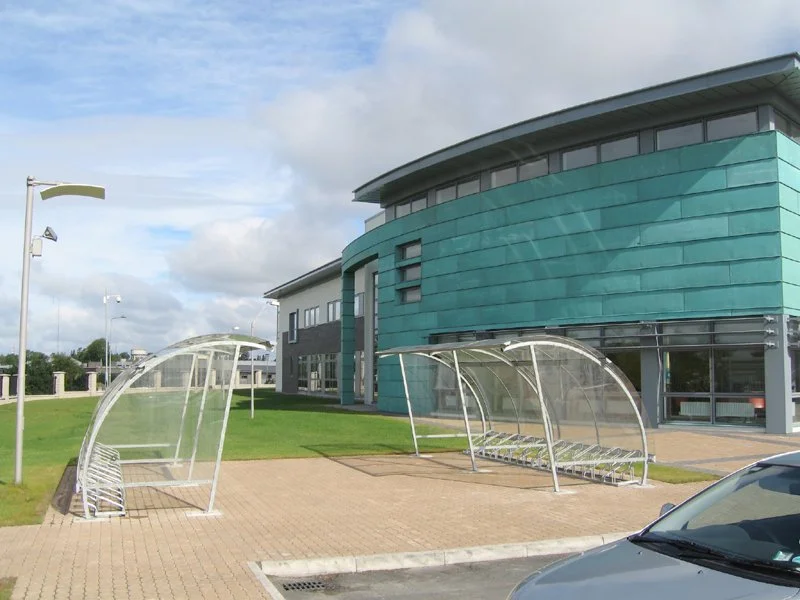 Modern building with curved roof and turquoise exterior, two glass-covered bike shelters in front, grassy area, and parking lot with a car in the foreground.