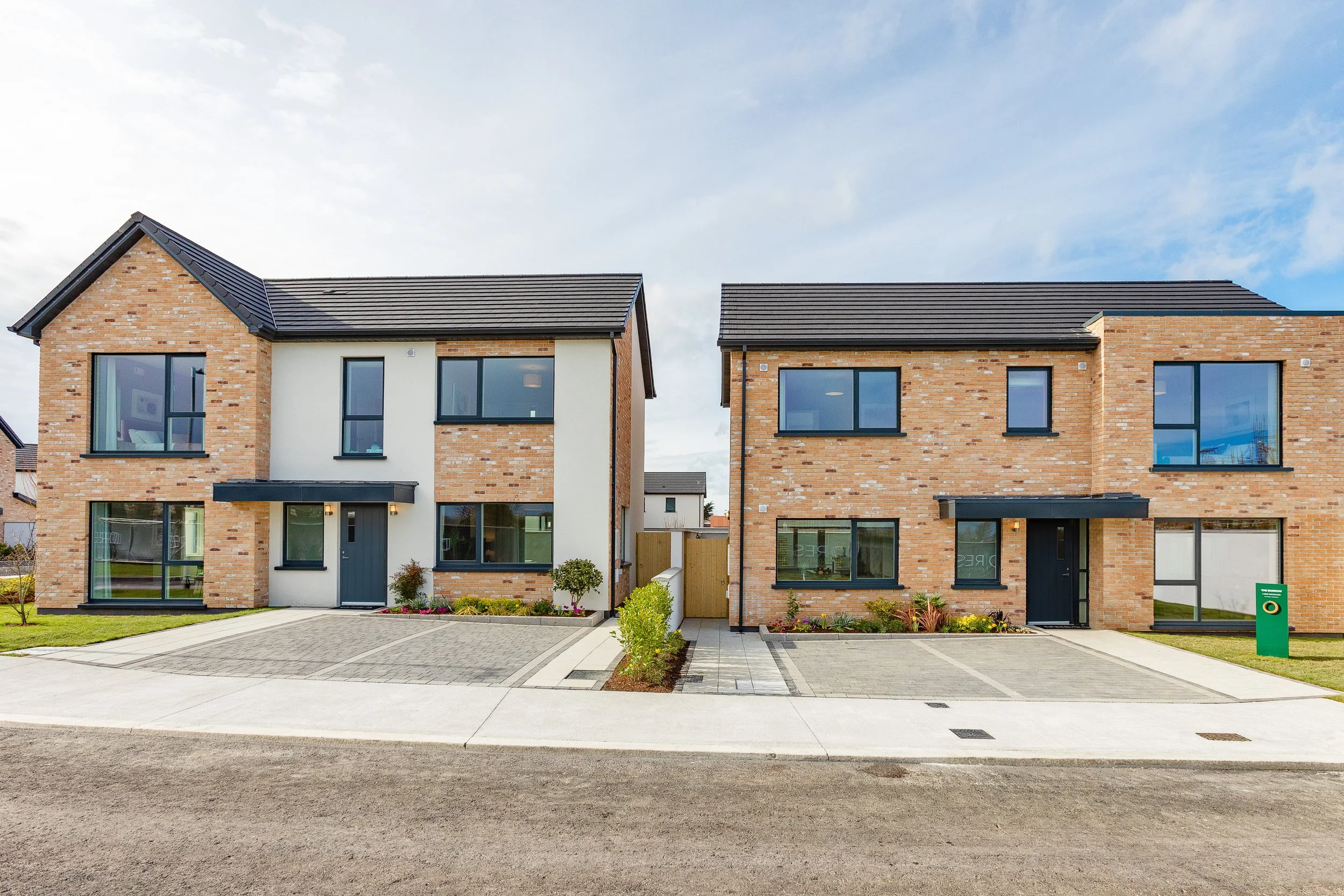 Two modern brick houses with dark gray roofs and black window frames on a residential street under a partly cloudy sky.