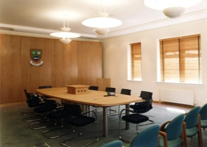 A conference room with a large wooden table surrounded by black chairs, and a few blue chairs on the side. The room has three windows with wooden blinds, wooden wall paneling, and white ceiling lights. There are some small items on the table, including a phone. An emblem or crest is mounted on the wooden wall.