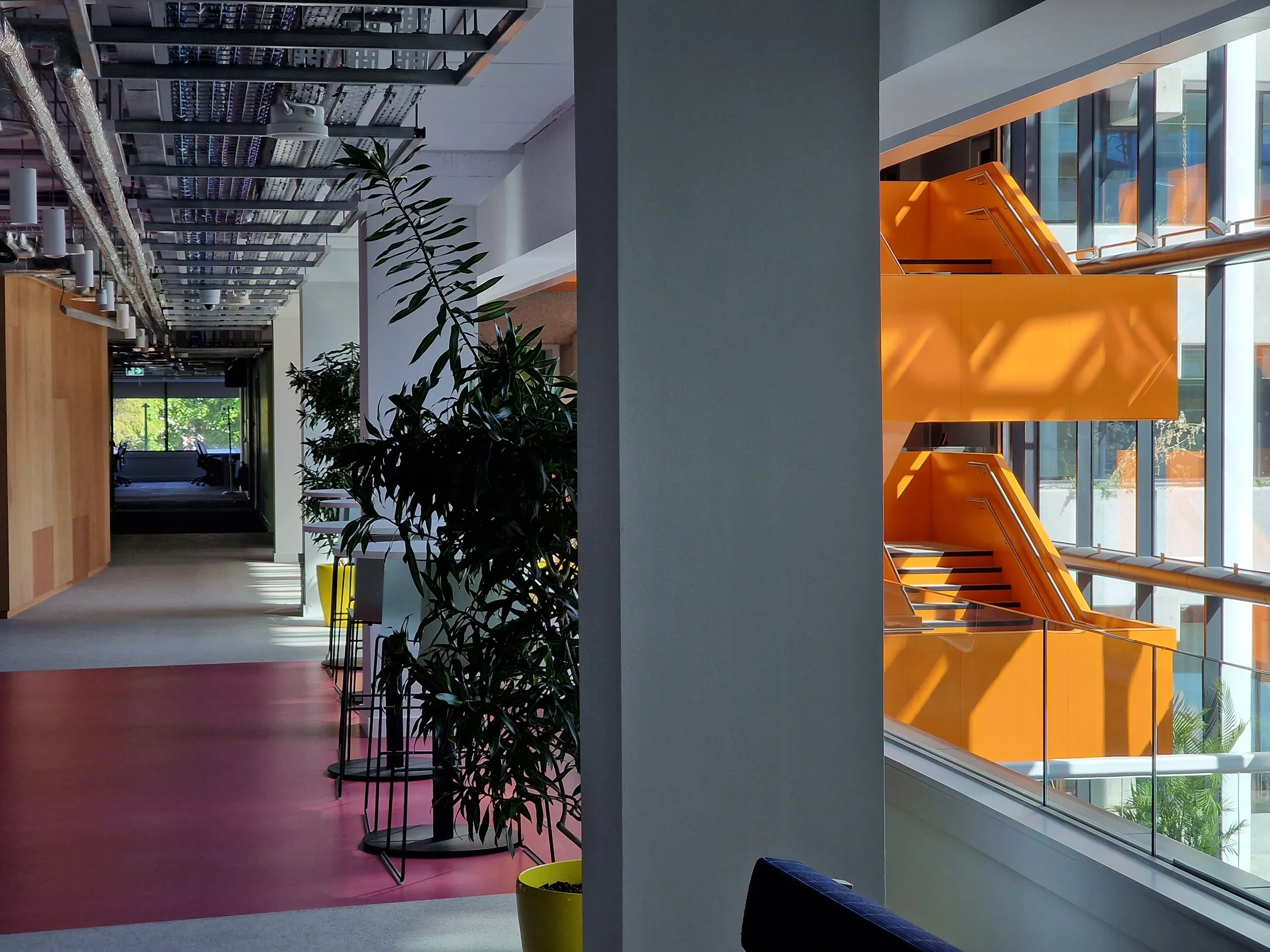 Modern office lobby with plants, a staircase with orange walls, large windows, polished floors, and seating areas.