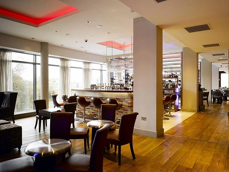 Modern hotel bar with stools, a high counter, and large windows with drapes, illuminated by red ceiling lights.