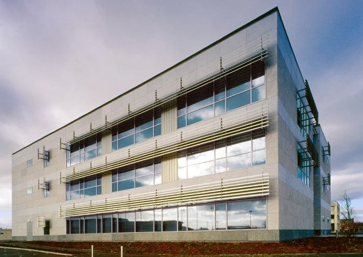 Modern office building with large glass windows and horizontal metal sunshades on a cloudy day.