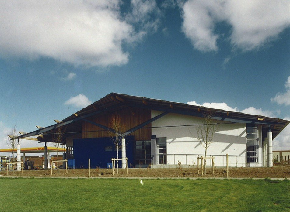 A modern building with a sloped roof, white and brown exterior, surrounded by a grassy area and young trees, under a blue sky with clouds.