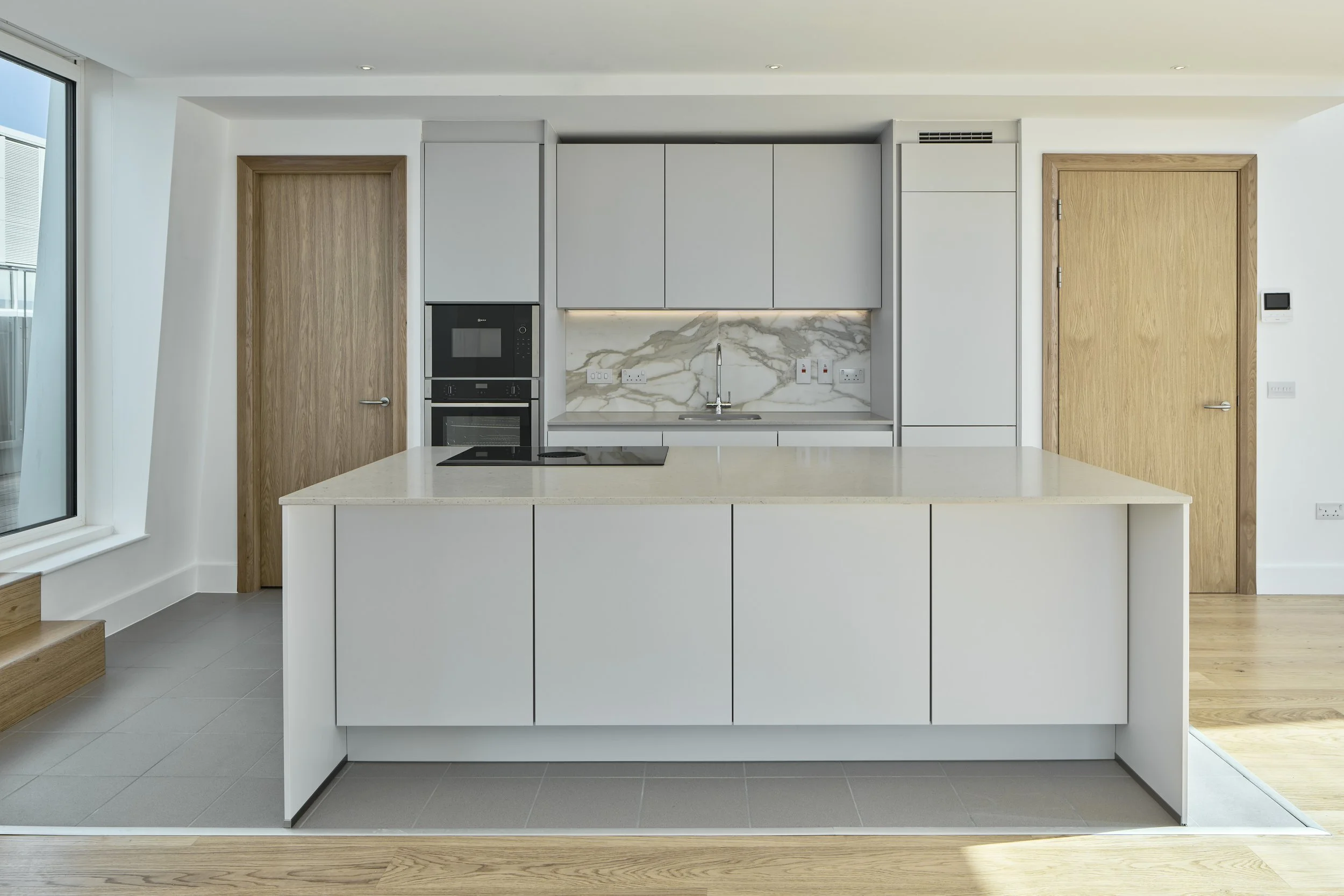Modern kitchen with white cabinetry, marble backsplash, wooden doors, island with light-colored countertop, and built-in appliances.