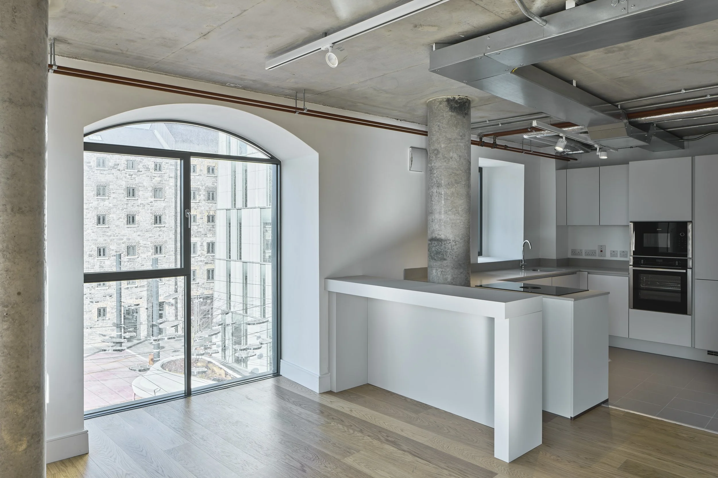 Modern kitchen in an open loft with large window, white cabinets, built-in oven, and a concrete pillar. Exposed ceiling ductwork and hardwood floors.