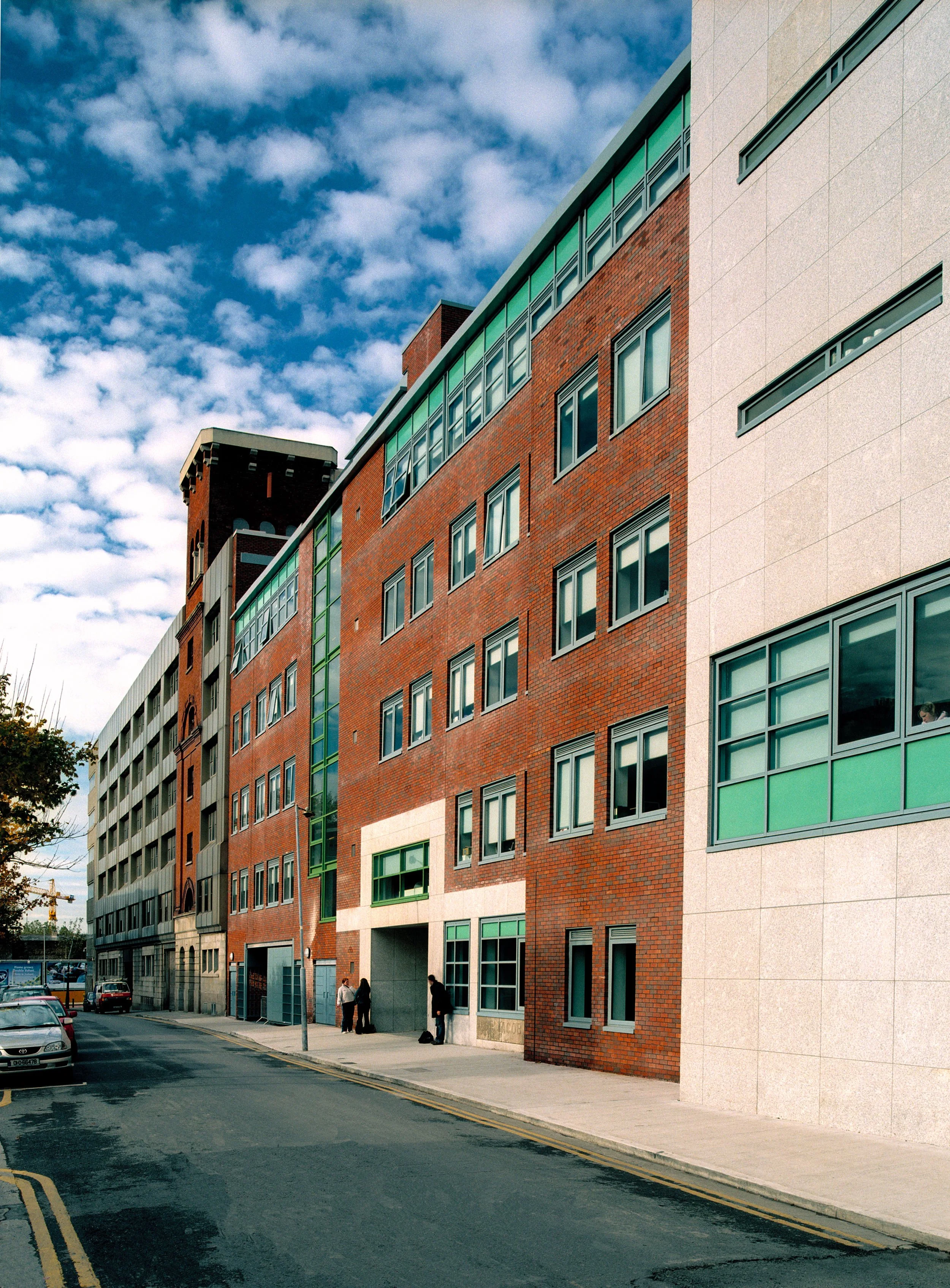 A city street view showing modern multi-story buildings with brick and concrete facades, several windows, and a few pedestrians walking along the sidewalk. Cars are parked along the street under a partly cloudy sky.