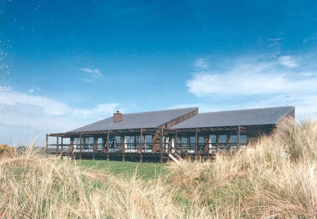 A large wooden house with a deck, situated in a grassy, dune-like area under a partly cloudy blue sky.