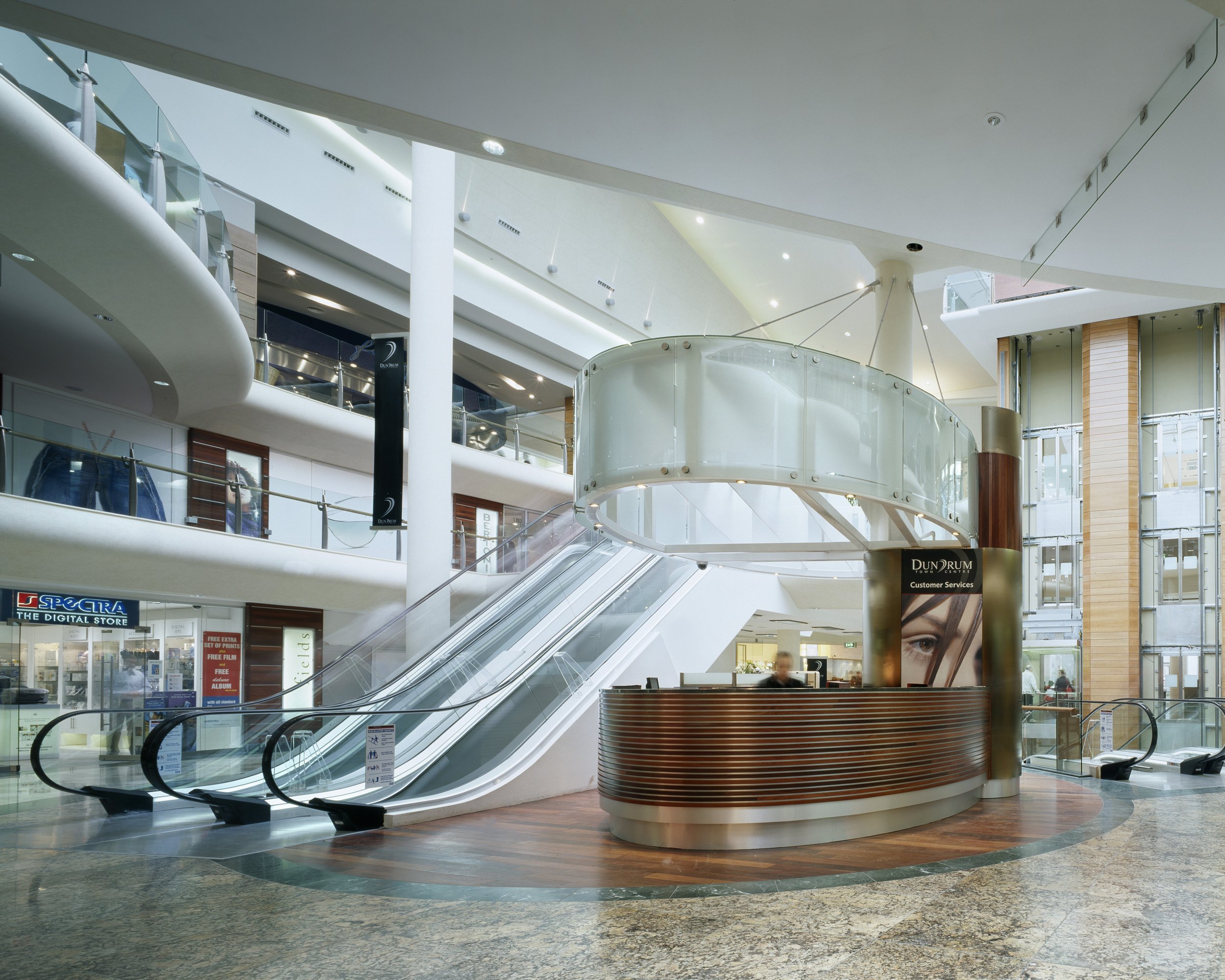 Interior view of a modern shopping mall with escalators, customer service desk, and multiple levels of stores and walkways.