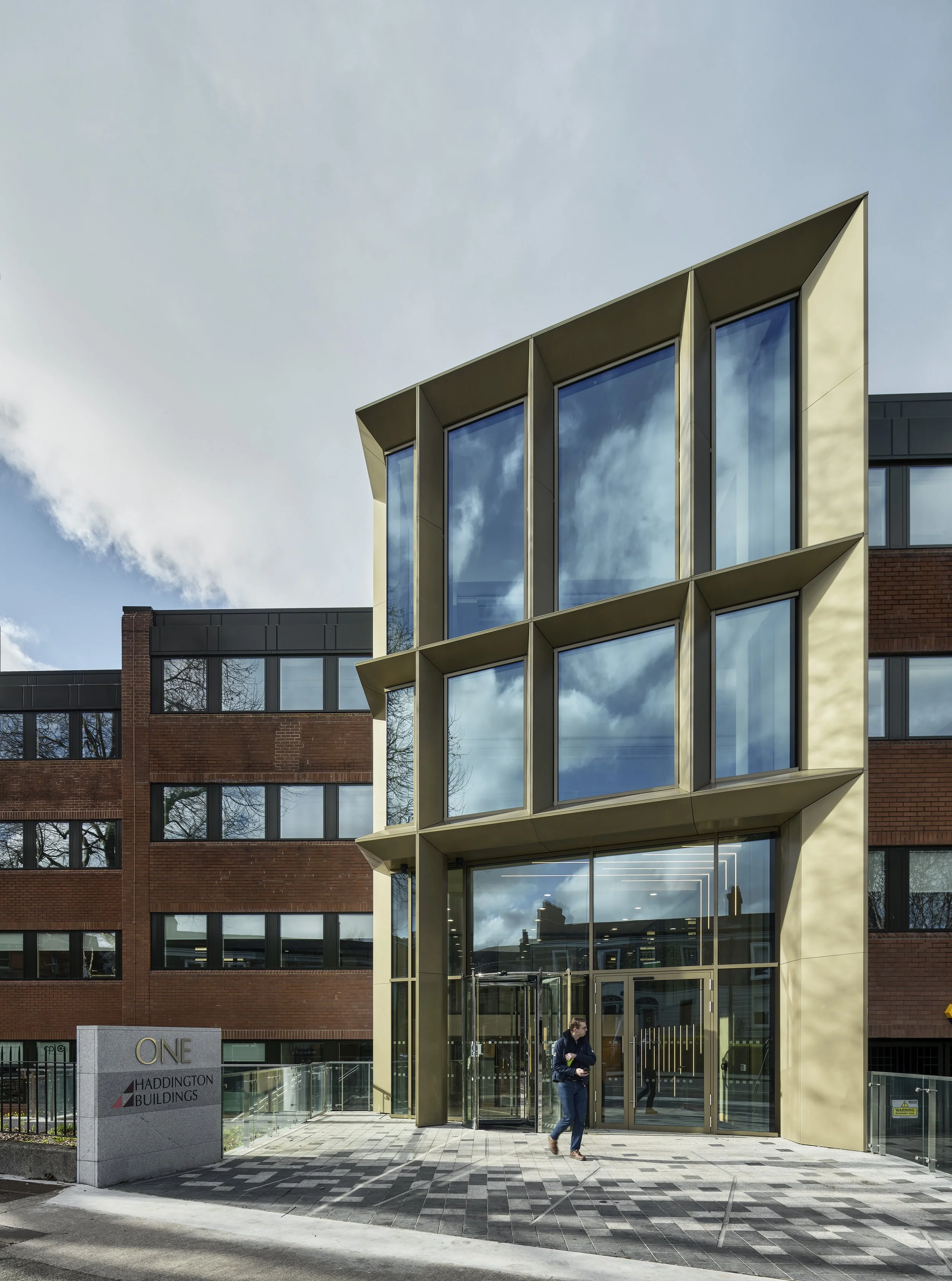 Modern glass and gold-colored building entrance with a person walking out, adjacent to a brick building, on a partly cloudy day.