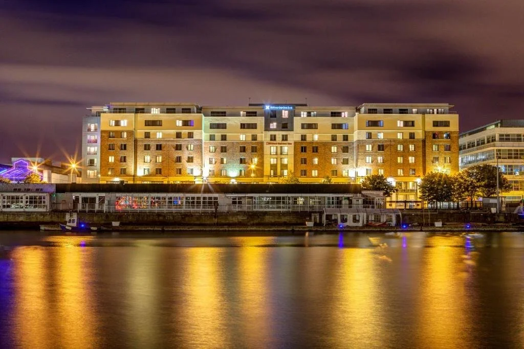 Nighttime view of a brightly lit apartment building across a river with reflections in the water.