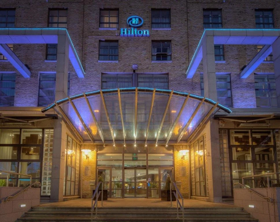 Night view of the entrance to a Hilton hotel with a blue Hilton sign above the door, glass doors, and a curved glass awning with lighting.