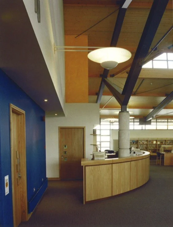 Interior of a modern building with wood and blue walls, a curved wooden reception desk, a large round ceiling light, and exposed beams on the ceiling.