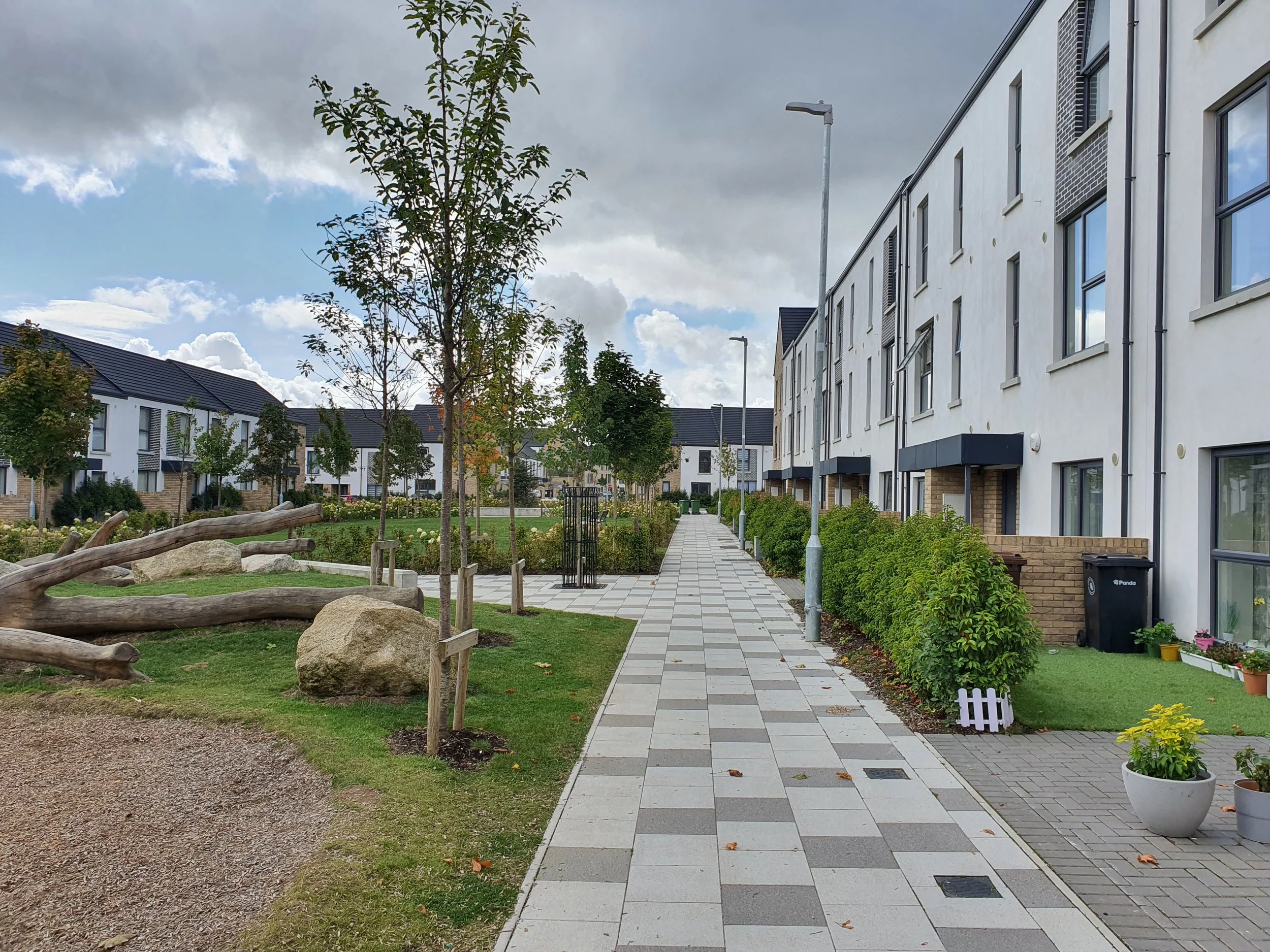 A residential street with modern white apartment buildings on the right and a landscaped park area with trees, rocks, and logs on the left. The sidewalk is paved with patterned tiles and is lined with bushes and streetlights, with a cloudy sky overhead.