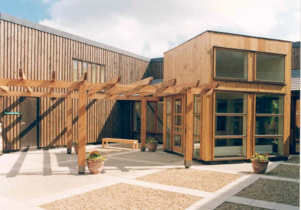 A modern building with wooden exterior and large windows, featuring a wooden pergola and potted plants in an outdoor courtyard.