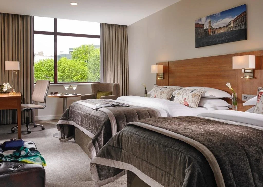 A hotel room with two beds, a large window with curtains showing green trees outside, a desk with a chair, and a sitting area with an armchair and a table.