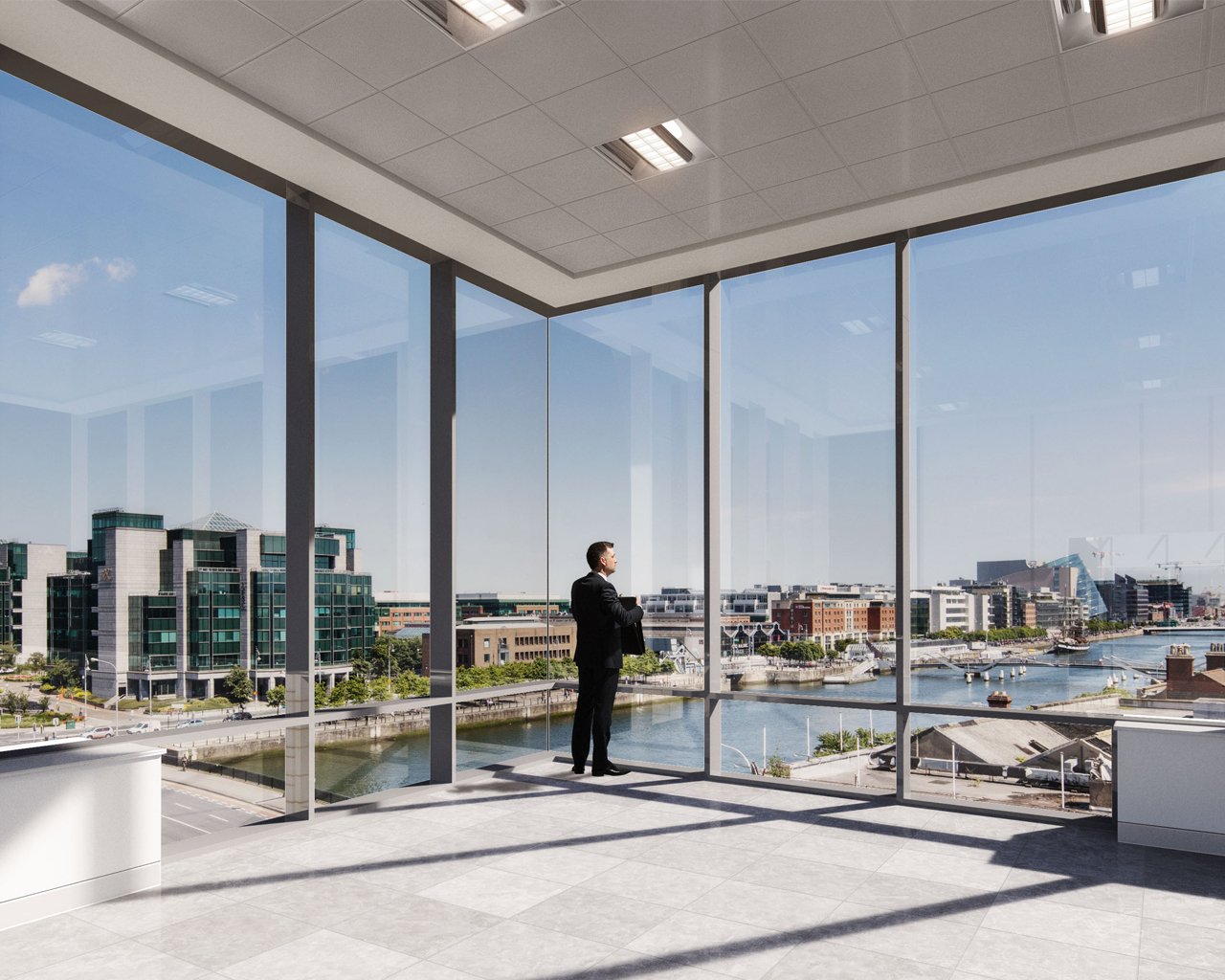 A man in a business suit standing inside a modern office with large floor-to-ceiling windows overlooking a cityscape with buildings, a river, and a clear blue sky.