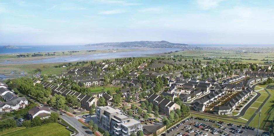 Aerial view of a residential neighborhood near a body of water, with houses, green spaces, and parking lots in the foreground and a distant shoreline and hills in the background.