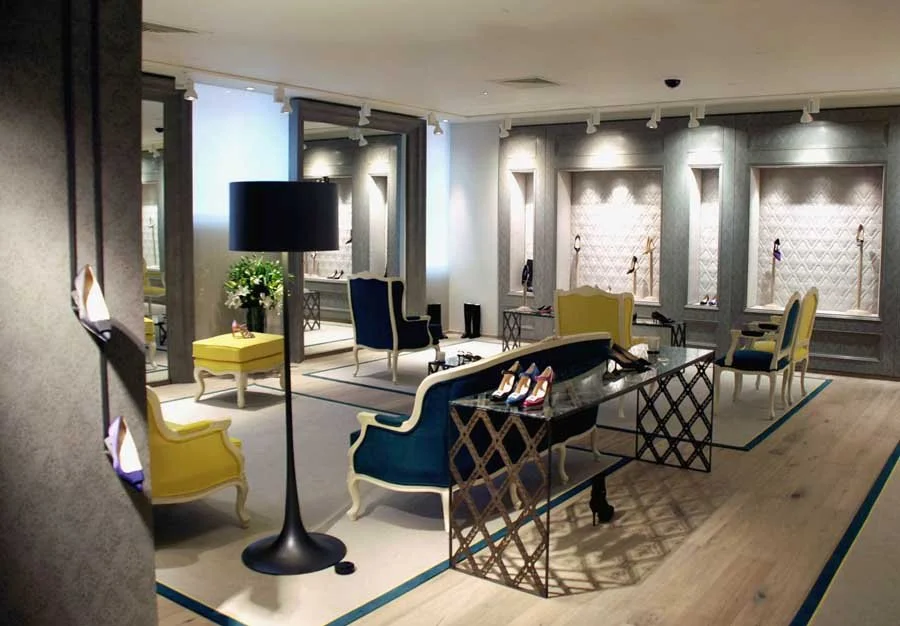 An elegant boutique with colorful chairs, a black table displaying shoes, and decorative niches on the wall with handbags, illuminated by spotlights.