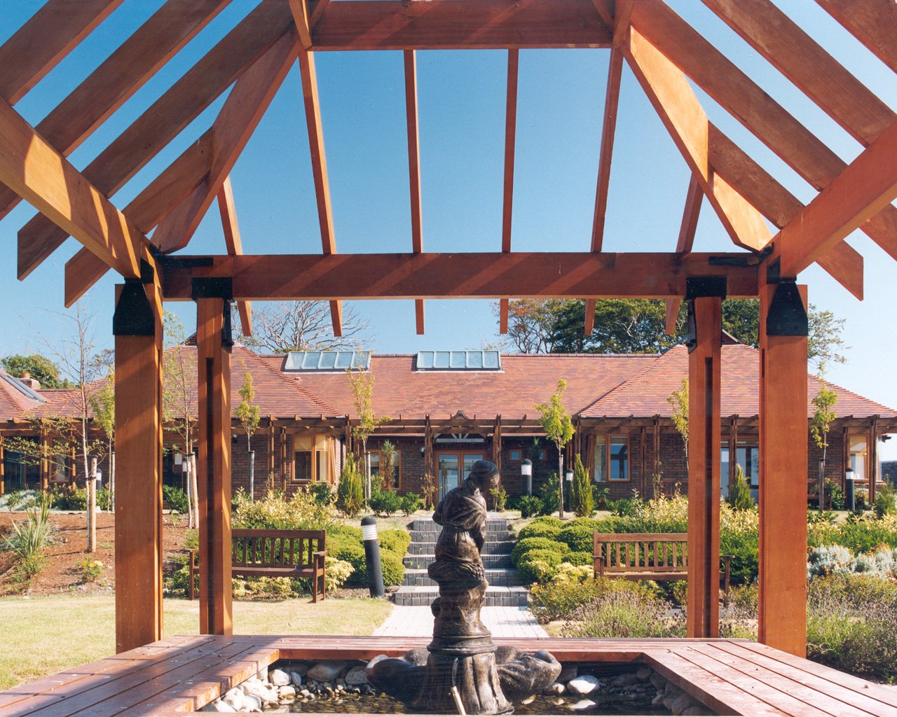 View from underneath a wooden pergola in a garden, with a fountain and statue in the center, surrounded by green bushes and trees, with a house in the background.