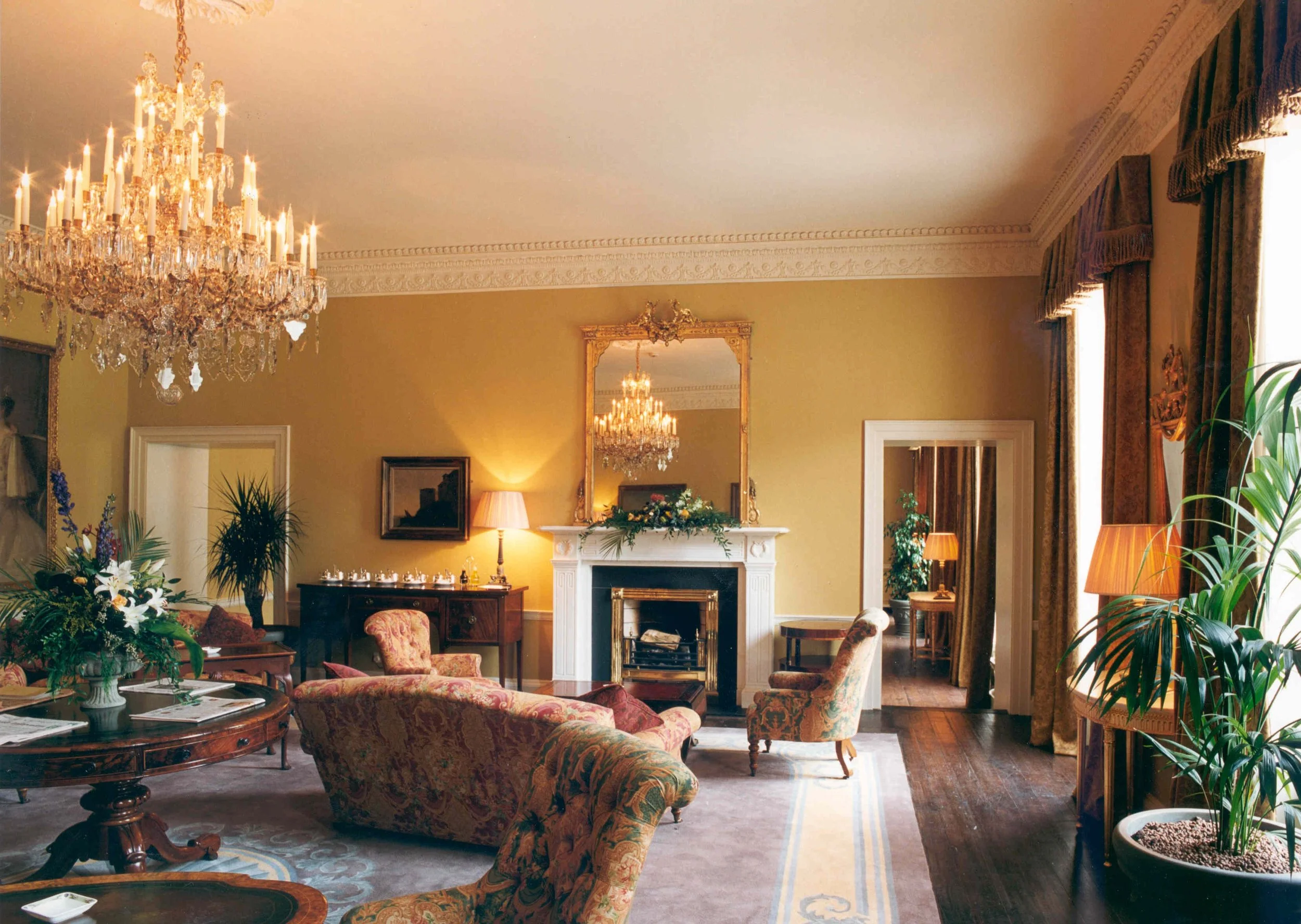 Elegant living room with chandelier, fireplace with mirror above, floral patterned couches, wooden chairs, potted plants, side tables with lamps, and large windows with curtains.