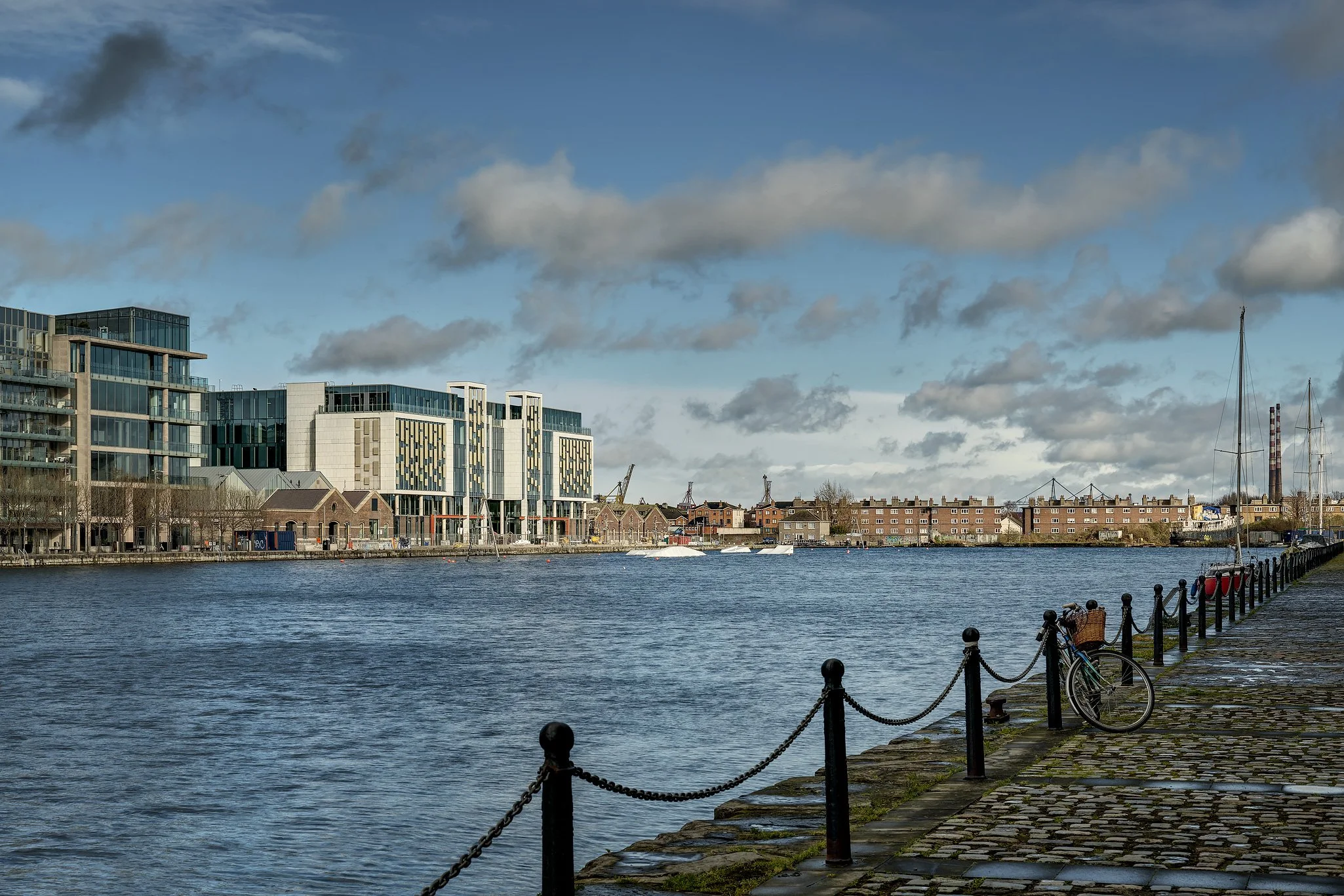 View of a river with modern buildings and houses along the shoreline, boats on the water, and a paved walkway with a bicycle and a chain-linked fence in the foreground under a partly cloudy sky.