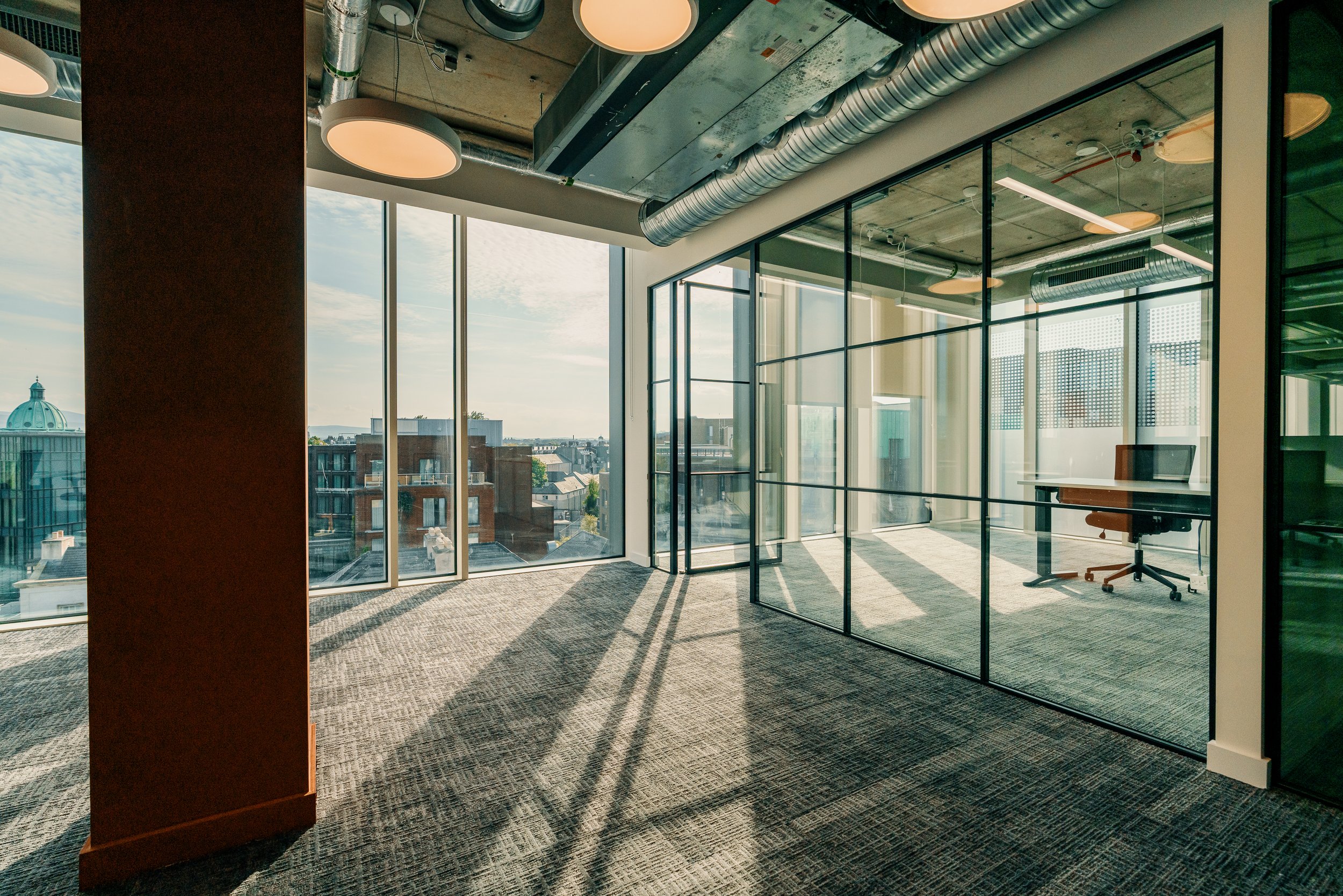 Empty office with large windows, glass walls, and office furniture, showing a cityscape view outside.
