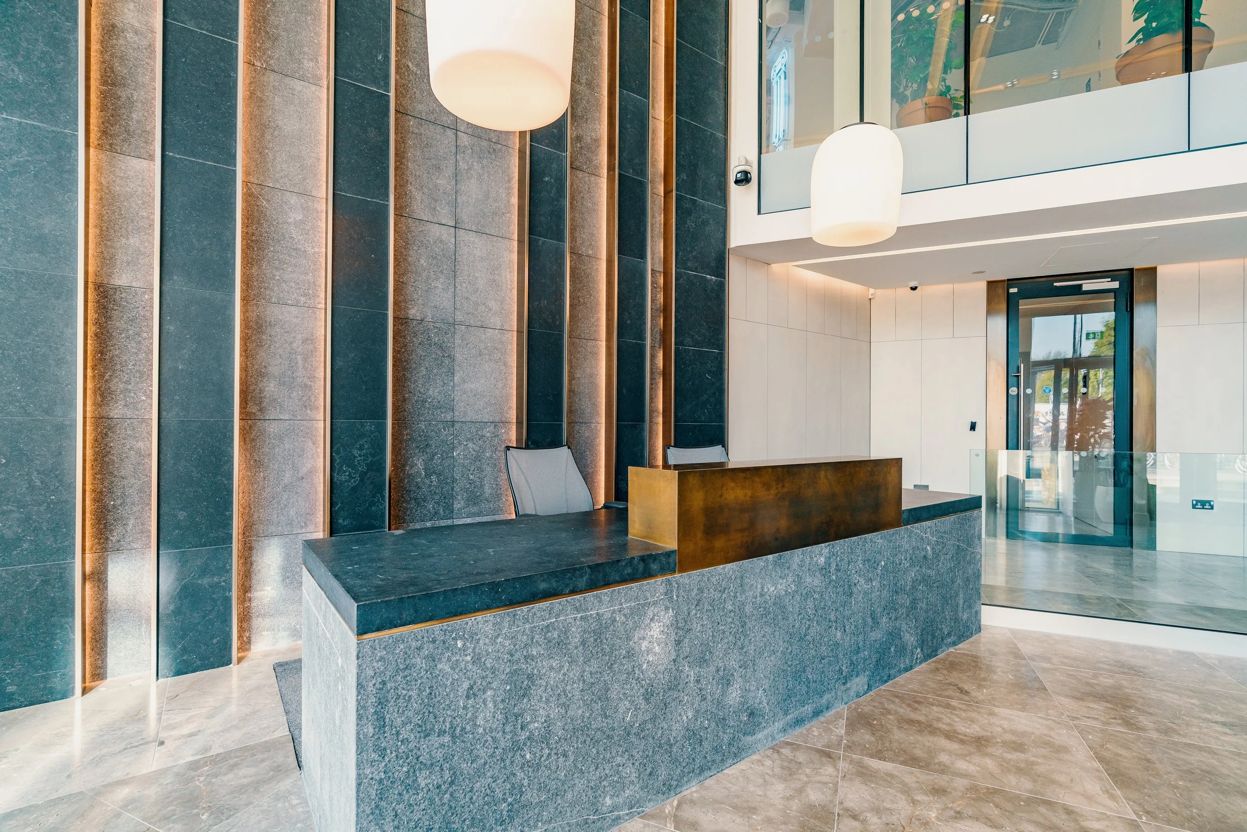 Modern hotel lobby reception desk with gray stone finishing, two sleek chairs behind the desk, large hanging white lamps, and a glass entrance door with a view of outside trees and street.
