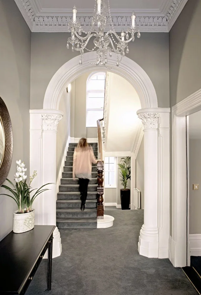 Interior of a house featuring a grand staircase with gray carpet, white ornate columns, a crystal chandelier, and a small hallway with potted plants.