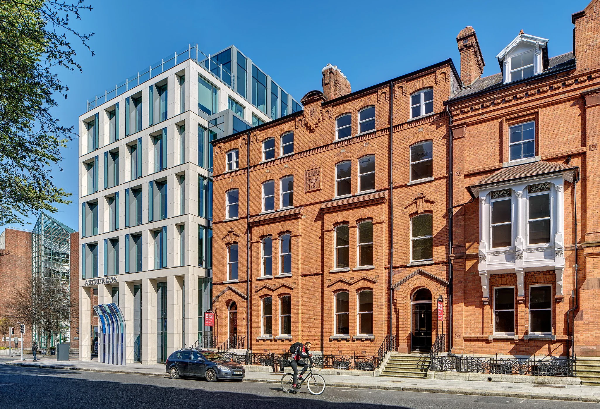 Modern white multi-story building with blue glass windows next to traditional red brick buildings on a city street with a cyclist and parked car.
