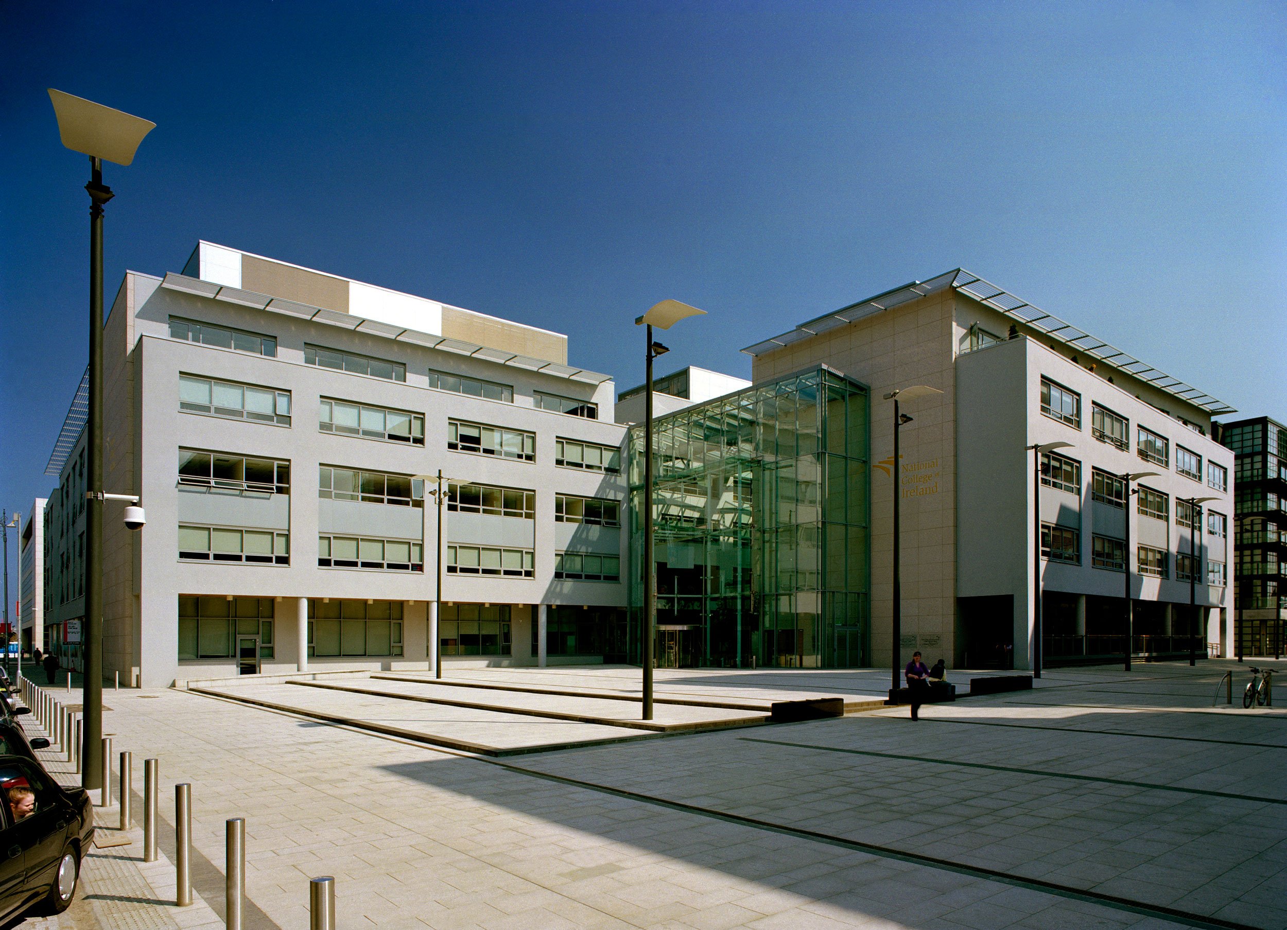 Modern building with a glass entrance, open plaza, street lamps, and a person sitting on a bench under a clear blue sky.