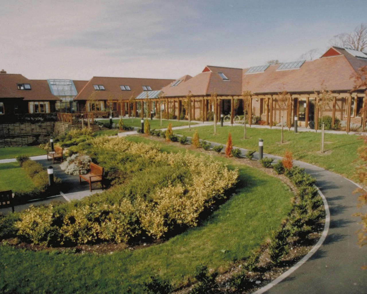 A landscaped courtyard with benches, a winding pathway, small trees, and residential buildings in the background under a partly cloudy sky.