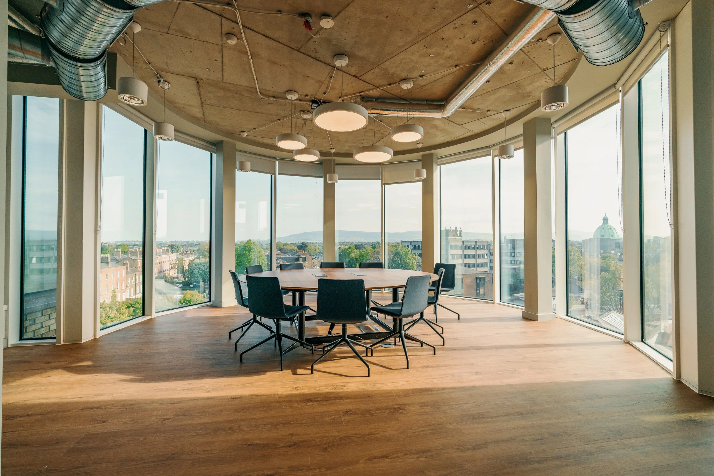 Modern conference room with large windows, round wooden table, and eight black chairs, overlooking city skyline with distant hill and dome building, under a ceiling with large circular light fixtures and exposed ductwork.