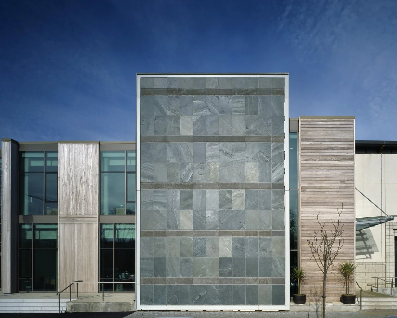 Modern building facade with large gray stone tiles, wood paneling, and glass windows against a clear blue sky.