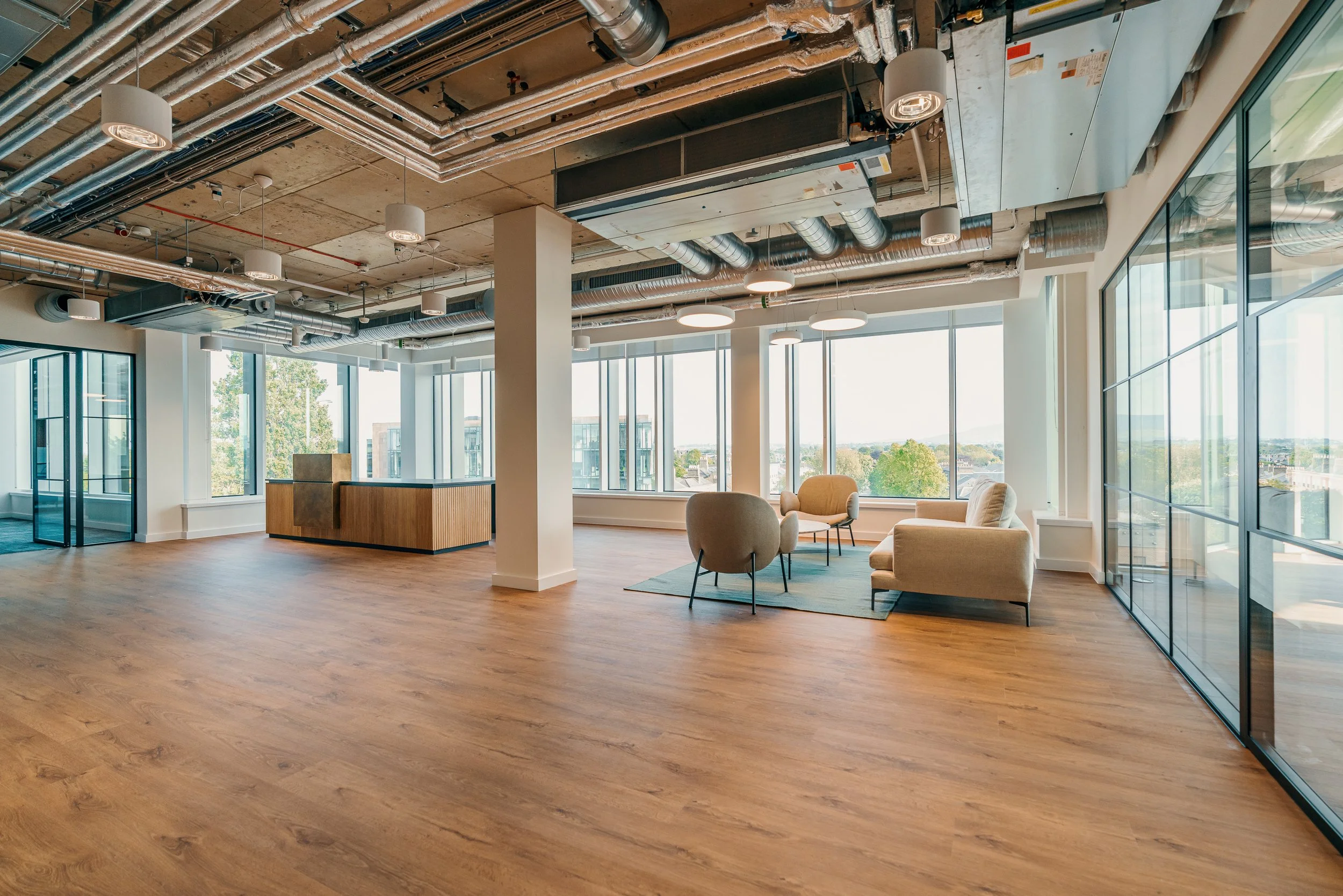Modern office lounge with large windows, wooden floors, beige sofas, armchairs, and a glass wall on the right.