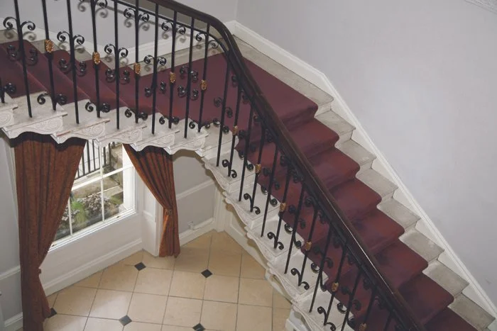 Interior view of a staircase with a wooden handrail and decorative metal balusters, with a window and curtains underneath.